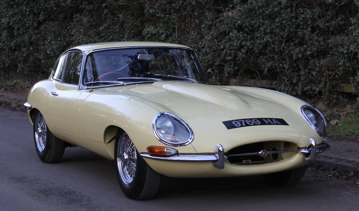 A Series 1 Jaguar E-Type fixed-head coupe in pale yellow parked on a quiet road